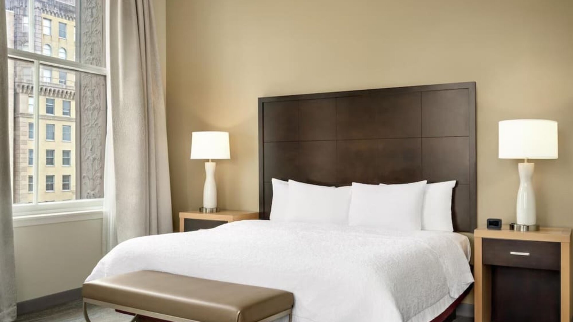Hotel guest room with a king bed, white bedding, dark wood headboard, bedside lamps, upholstered bench, and tall window overlooking neighboring city buildings.