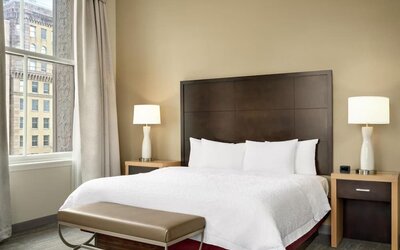 Hotel guest room with a king bed, white bedding, dark wood headboard, bedside lamps, upholstered bench, and tall window overlooking neighboring city buildings.