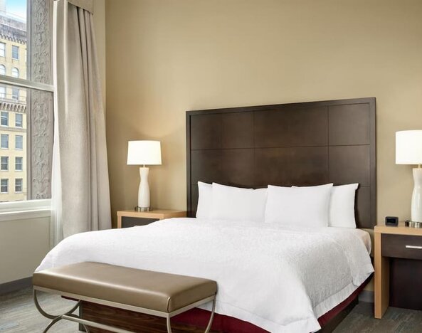 Hotel guest room with a king bed, white bedding, dark wood headboard, bedside lamps, upholstered bench, and tall window overlooking neighboring city buildings.