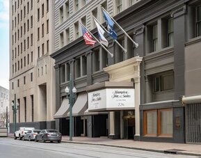 Street-level view of a multi-story urban hotel with a dark gray façade, columned entrance canopy, American and other flags above the doorway, and parked cars along the curb.