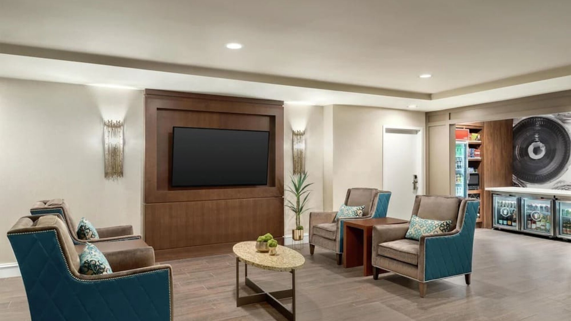 Comfortable hotel lounge with teal and gray upholstered armchairs, patterned cushions, oval coffee table, wall-mounted flat-screen TV, decorative lighting, and access to the adjacent convenience shop.