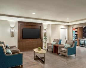 Comfortable hotel lounge with teal and gray upholstered armchairs, patterned cushions, oval coffee table, wall-mounted flat-screen TV, decorative lighting, and access to the adjacent convenience shop.