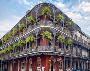 Historic New Orleans corner building with red exterior walls, ornate wrought-iron balconies on multiple levels, abundant hanging ferns, ground-floor storefronts, bicycles, and pedestrians along the street.