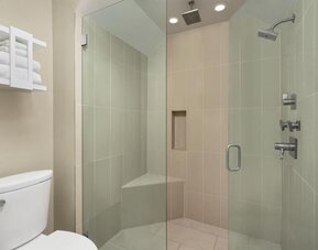 Modern hotel bathroom with a glass-enclosed walk-in shower, tiled walls, rainfall-style showerhead, built-in corner seat and wall niche, toilet, and open shelving with folded towels.