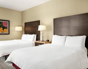 Hotel guest room with two beds dressed in white linens, tall dark wood headboards, shared nightstand with lamp, beige walls, and colorful floral artwork above one bed.
