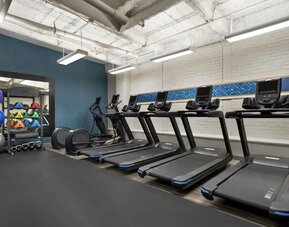 Modern hotel fitness center with five treadmills, elliptical equipment, kettlebells, medicine balls, stability balls, white brick walls, and suspended rectangular lighting.