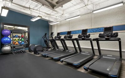 Modern hotel fitness center with five treadmills, elliptical equipment, kettlebells, medicine balls, stability balls, white brick walls, and suspended rectangular lighting.