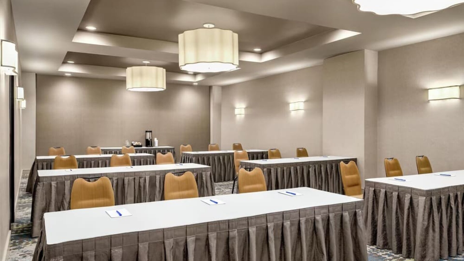 Hotel meeting room arranged classroom-style with long white tables, tan chairs, patterned carpet, large pendant lights, and notepads with pens at each place.