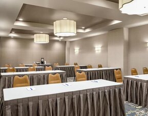 Hotel meeting room arranged classroom-style with long white tables, tan chairs, patterned carpet, large pendant lights, and notepads with pens at each place.