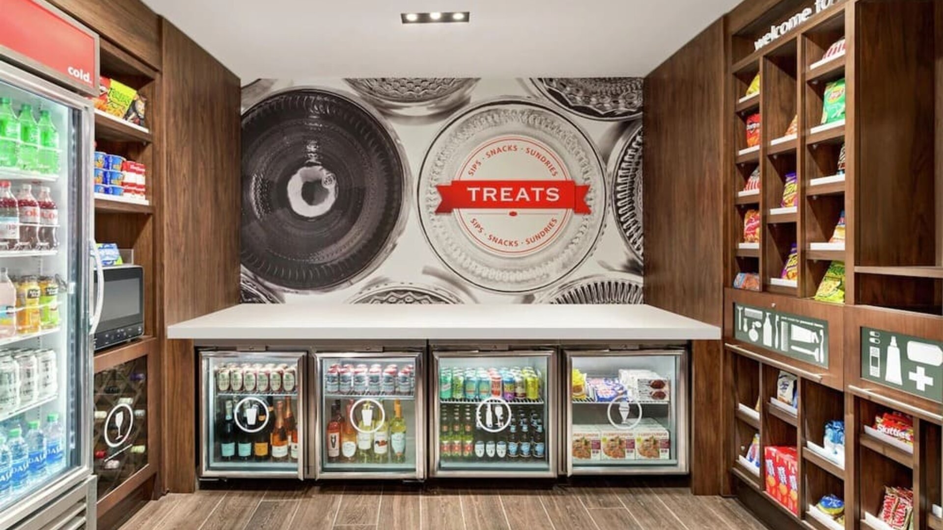 Hotel convenience shop with wooden shelving stocked with snacks, refrigerated cases containing bottled and canned drinks, and a central counter beneath a large “Treats” wall graphic.
