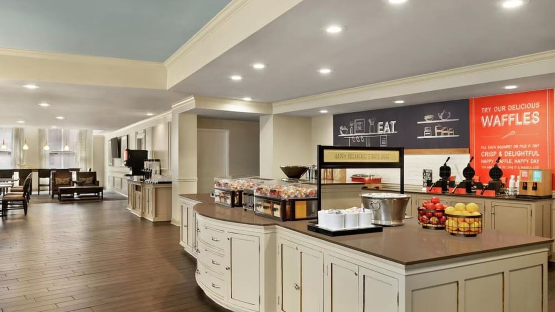 Spacious hotel dining and breakfast area with a large buffet island, fruit displays, beverage stations, waffle-making area, wood-look flooring, and lounge seating in the background.