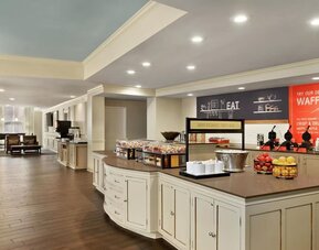 Spacious hotel dining and breakfast area with a large buffet island, fruit displays, beverage stations, waffle-making area, wood-look flooring, and lounge seating in the background.