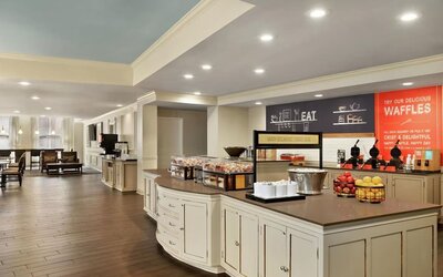 Spacious hotel dining and breakfast area with a large buffet island, fruit displays, beverage stations, waffle-making area, wood-look flooring, and lounge seating in the background.