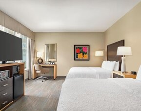 Spacious hotel guest room with two beds dressed in white linens, dark wood headboards, bedside lamps, desk and chair, flat-screen TV, microwave, mini refrigerator, and colorful framed artwork.