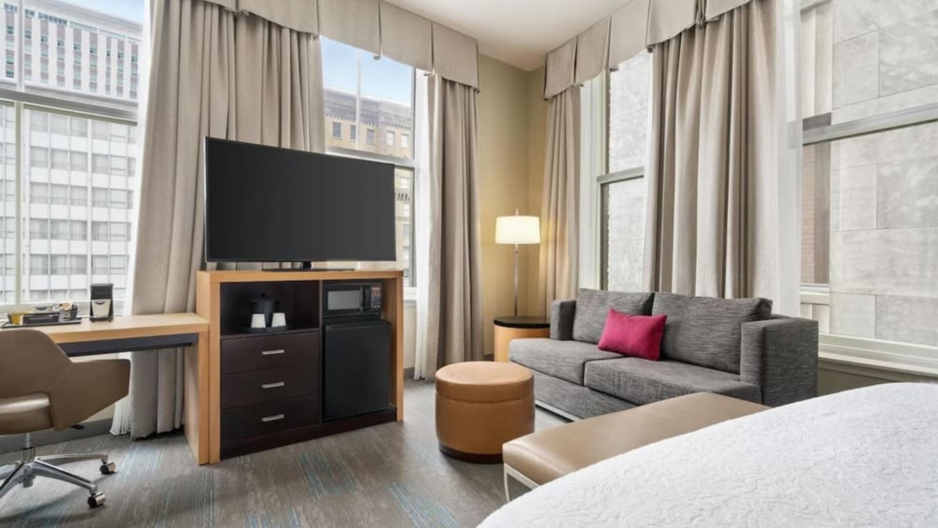 Bright corner hotel room with large windows, gray sofa with pink accent pillow, desk and chair, ottoman, flat-screen TV, microwave, mini refrigerator, and part of a bed visible.