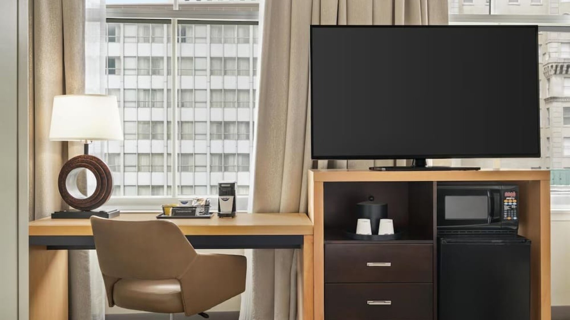 Guest room workspace with a desk and swivel chair beside a large window, flat-screen TV, microwave, mini refrigerator, storage drawers, coffee supplies, and decorative table lamp.