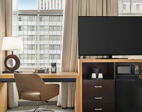Guest room workspace with a desk and swivel chair beside a large window, flat-screen TV, microwave, mini refrigerator, storage drawers, coffee supplies, and decorative table lamp.