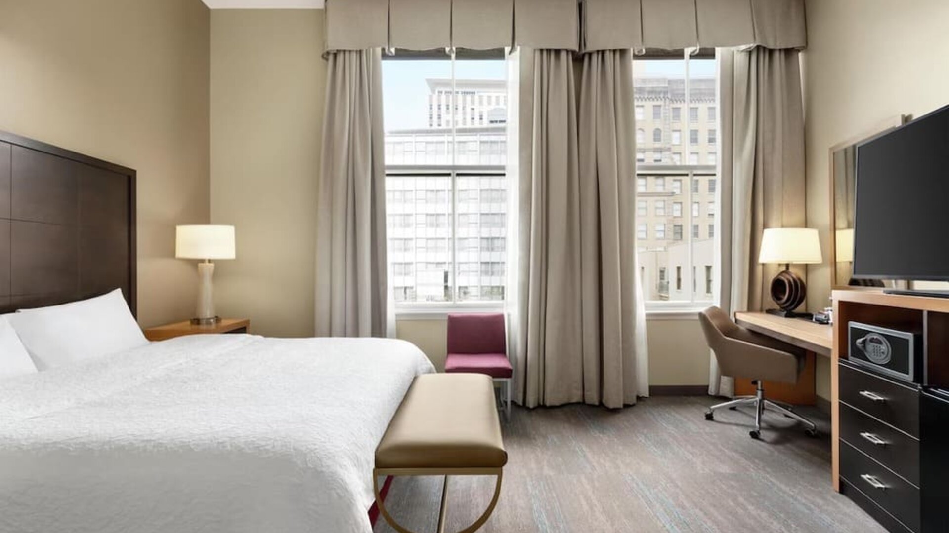 Spacious hotel room with a king bed, white linens, upholstered bench, desk and swivel chair, flat-screen TV, storage unit, tall windows, and a red accent chair overlooking the city.