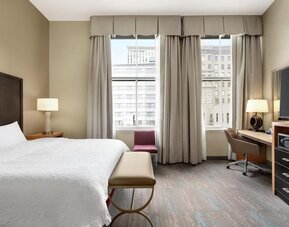 Spacious hotel room with a king bed, white linens, upholstered bench, desk and swivel chair, flat-screen TV, storage unit, tall windows, and a red accent chair overlooking the city.