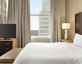 Hotel guest room with a king bed, white bedding, dark wood headboard, bedside lamp, flat-screen TV on a dresser, upholstered bench, and tall window overlooking surrounding city buildings.