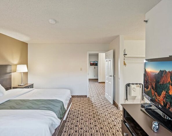 Spacious king guest room featuring a king bed, flat-screen television, storage space, and patterned carpeting at Coratel Plus Suites Wichita West Airport.