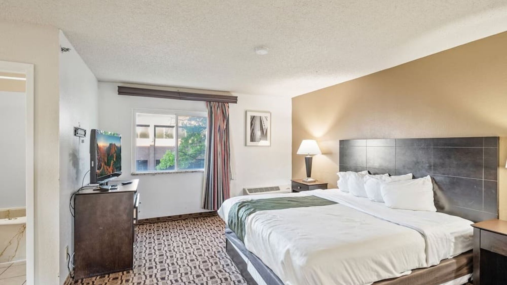 Bright king guest room featuring a king bed, large window providing natural light, flat-screen television, and bedside lighting at Coratel Plus Suites Wichita West Airport.