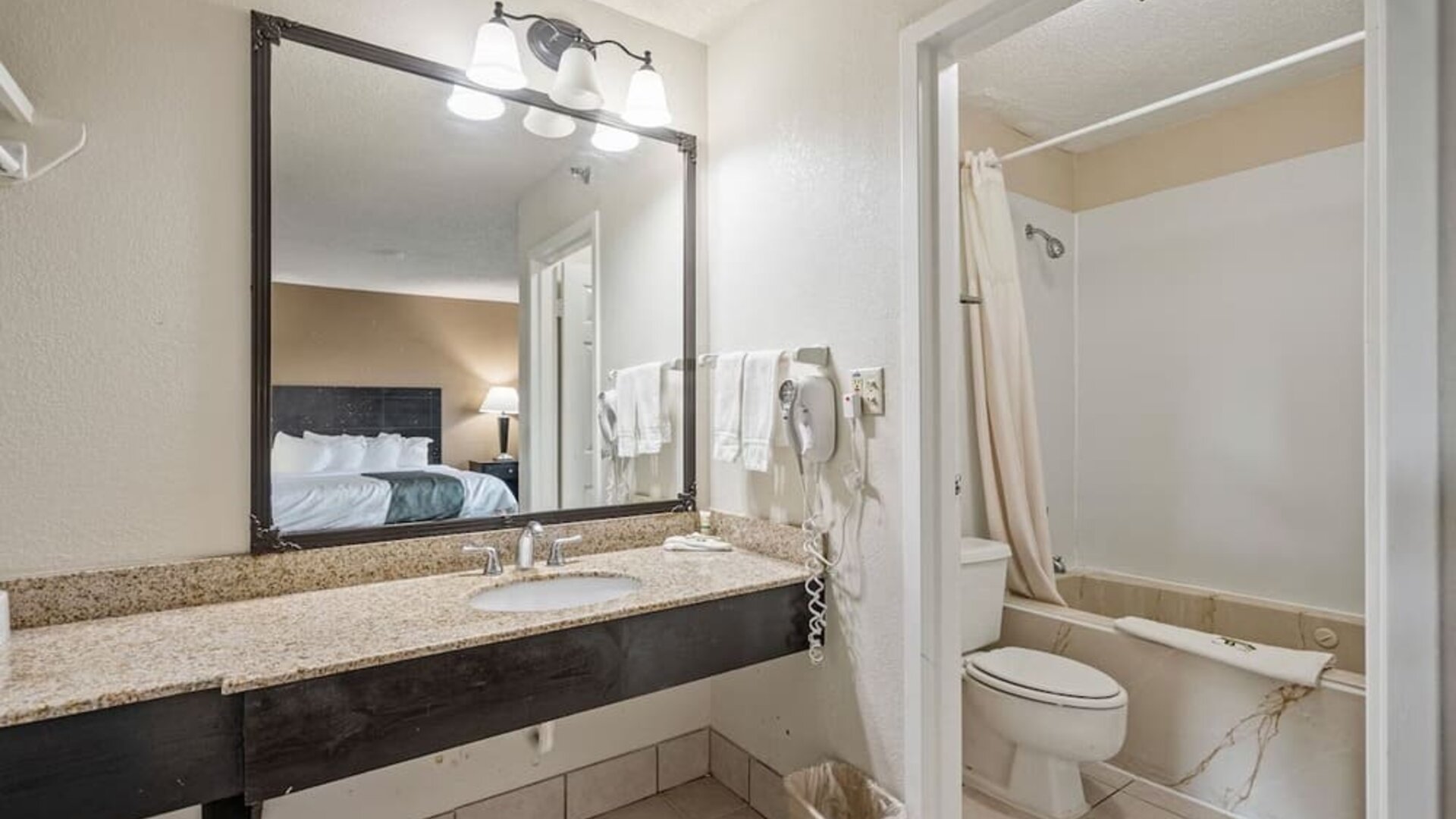 Guest bathroom featuring a granite vanity, large mirror, bathtub and shower combination, and fresh towels at Coratel Plus Suites Wichita West Airport.