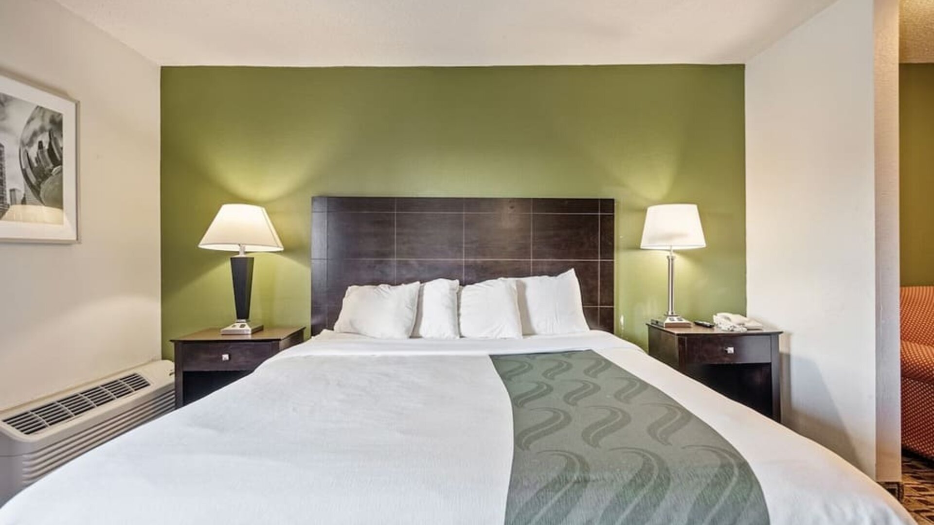 King guest room featuring a comfortable king bed, green accent wall, bedside tables, and warm lighting at Coratel Plus Suites Wichita West Airport.