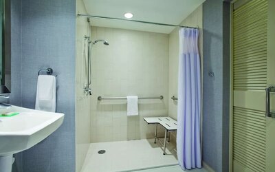 Accessible guest bathroom featuring a roll-in shower, grab bars, fold-down shower seat, and pedestal sink at Hyatt Place San Jose Downtown.