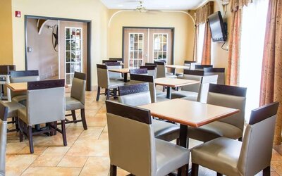 Bright hotel dining area with multiple tables, chairs, television, and large windows at Comfort Suites San Antonio SeaWorld - Lackland AFB.