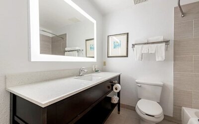 Guest bathroom with illuminated mirror, vanity, toilet, towels, and tiled bath area at SureStay by Best Western Lewisville Dallas.