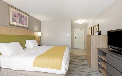 King guest room with white bedding, yellow bed runner, green upholstered headboard, television, dresser, and storage cabinet at SureStay by Best Western Lewisville Dallas.