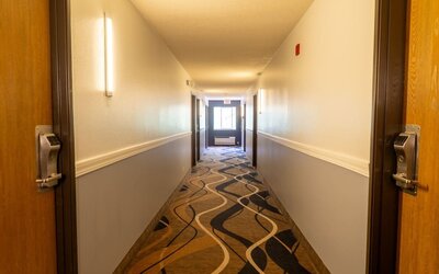 Hotel corridor with patterned carpeting, guest room doors, wall lighting, and window at the end of the hallway at Coratel Inn & Suites By Jasper Hastings.