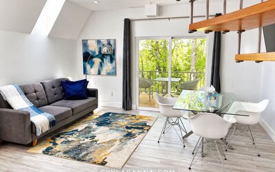 Open-plan cabin living and dining area at SKYLARANNA Resort & Spa, featuring a sofa, glass dining table, contemporary artwork, spiral staircase, and doors opening onto a private balcony.