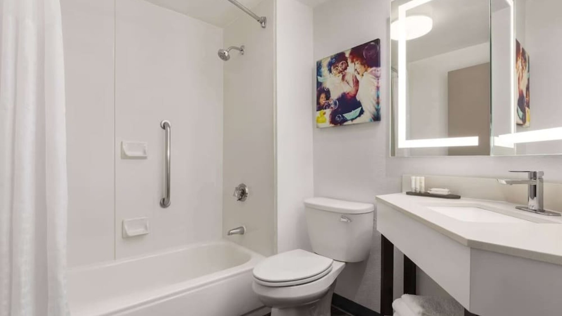 Modern guest bathroom featuring a shower and bathtub combination, illuminated mirror, vanity, fresh towels, and accessibility grab bar at La Quinta Inn & Suites By Wyndham Cleveland Airport West.