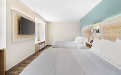Spacious modern guest room with two beds, white bedding, teal accent wall, wood flooring, and wall-mounted television.