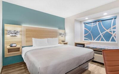 Modern king guest room with white bedding, teal accent wall, work desk, mini fridge, microwave, and window seating area.