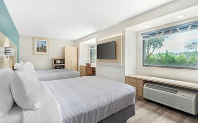 Bright guest room with two beds, large window overlooking greenery, wall-mounted TV, work desk, and air-conditioning unit. 
