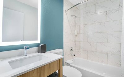 Bright hotel bathroom with illuminated mirror, modern vanity, toilet, bathtub, shower, and white marble-look tiled walls.