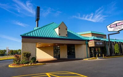 Front exterior of the hotel and City Café under a bright blue sky, featuring a teal metal roof, covered entrance, landscaped shrubs, paved driveway, and City Café signage.