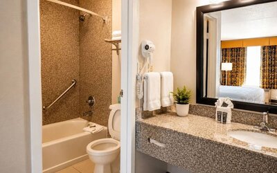 Hotel bathroom featuring a granite vanity with sink and large framed mirror, white towels, wall-mounted hair dryer, toilet, and bathtub with shower and grab bar.