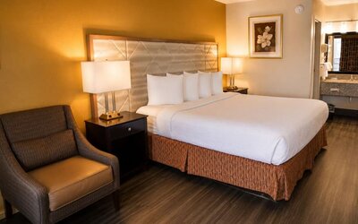 Spacious hotel room with a king bed, white bedding, padded headboard, mustard-gold accent wall, bedside lamps, armchair, wood flooring, and open vanity area.