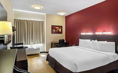 Modern hotel guest room with a king-size bed against a red accent wall, white linens, desk and chair, curtained window, air-conditioning unit, and dark wood furnishings.