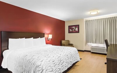 Spacious hotel room with a king-size bed, crisp white bedding, dark wood headboard, red accent wall, armchair, curtained window, and air-conditioning unit.