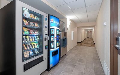 Hotel hallway with illuminated snack and Pepsi beverage vending machines, an ice machine, tiled flooring, beige walls, and guest room corridor extending into the distance.