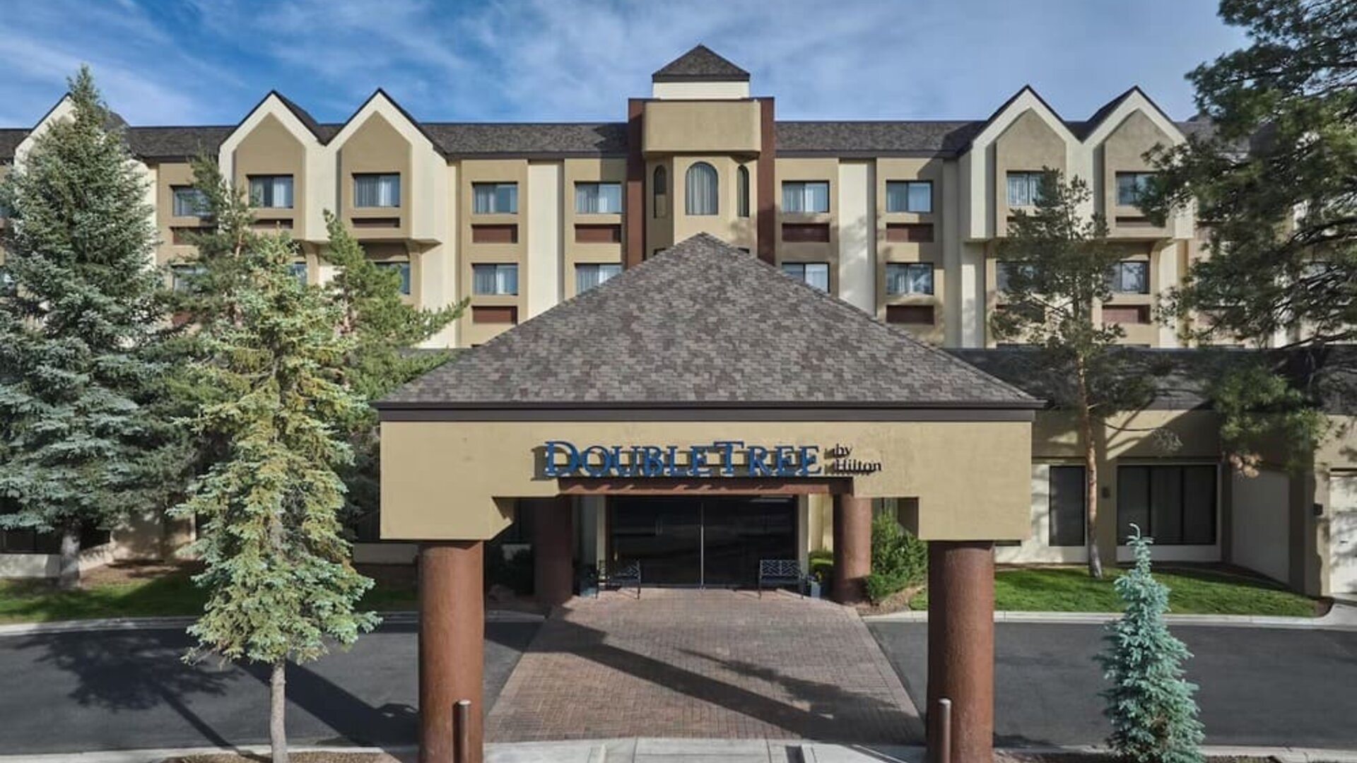 Daytime exterior view of the multi-story DoubleTree by Hilton hotel, featuring a covered entrance with hotel signage, beige facade, peaked rooflines, mature evergreen trees, and a paved driveway.