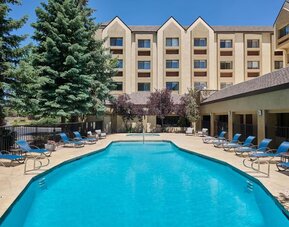 Large rectangular outdoor swimming pool surrounded by a sunny patio with blue lounge chairs, tables, mature evergreen trees, and the multi-story hotel building in the background.