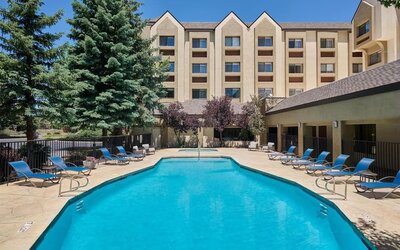Large rectangular outdoor swimming pool surrounded by a sunny patio with blue lounge chairs, tables, mature evergreen trees, and the multi-story hotel building in the background.