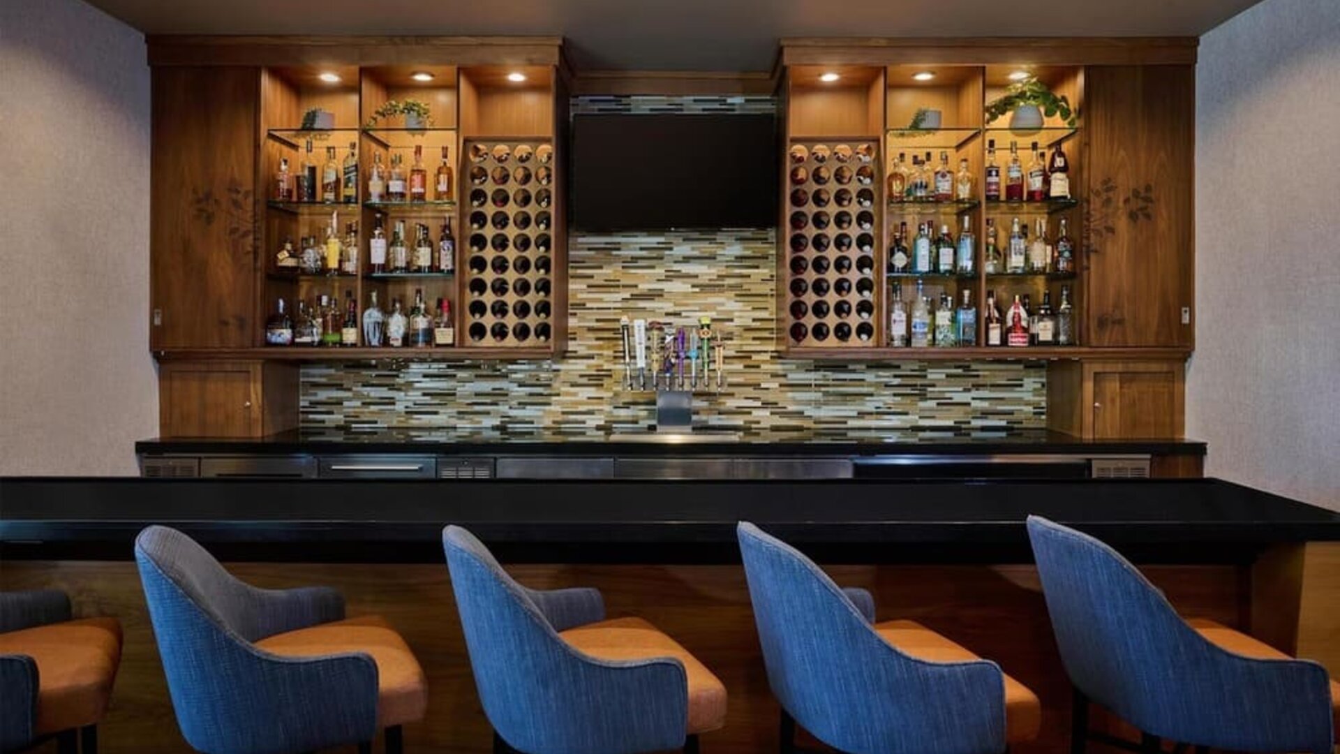 Contemporary hotel bar with a long black counter, blue upholstered bar stools, illuminated wooden shelving stocked with bottles, built-in wine racks, beer taps, and a wall-mounted television.