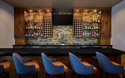 Contemporary hotel bar with a long black counter, blue upholstered bar stools, illuminated wooden shelving stocked with bottles, built-in wine racks, beer taps, and a wall-mounted television.